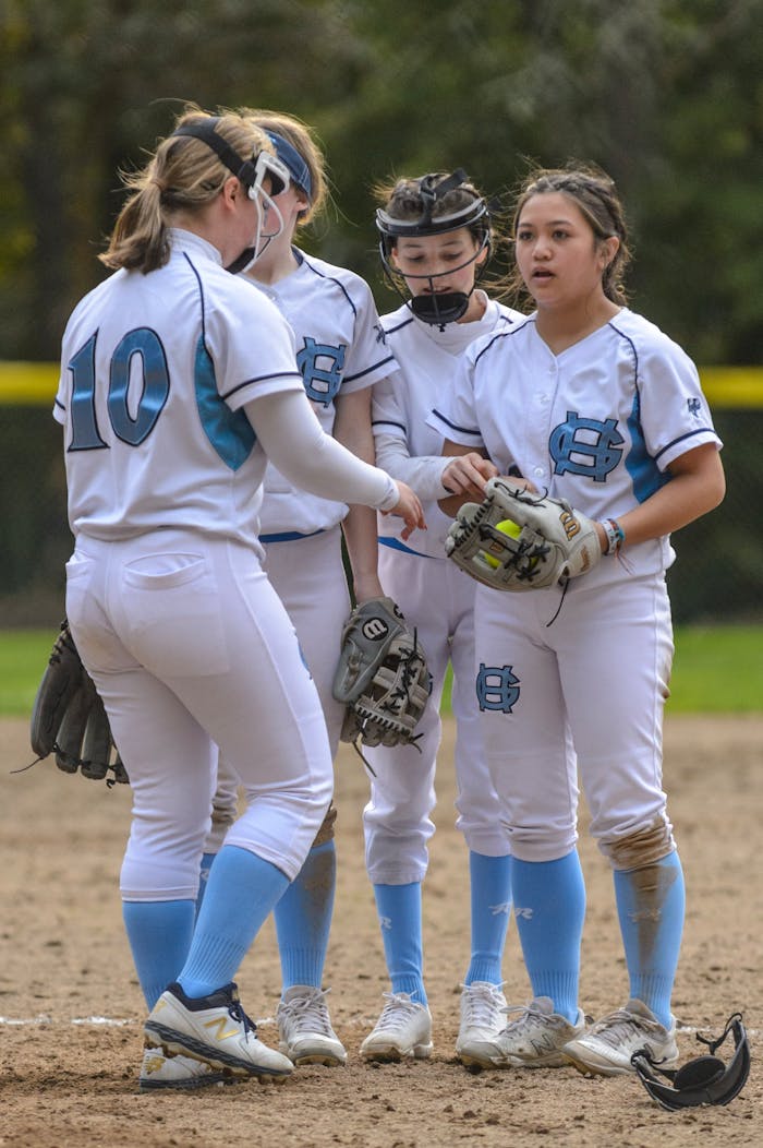 Four female softball players in uniform having a strategic discussion on the field during a daylight game.